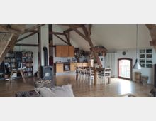 Geräumiger Dachbodenraum mit Holzsichtbalken, einer offenen Küche mit Holzmöbeln und Esstisch, einem Holzofen in der Mitte, Bücherregalen auf der linken Seite und einem großen Fenster mit Rundbogen auf der rechten Seite. Der Holzboden und die gemütliche Einrichtung verleihen dem Raum eine rustikale Atmosphäre.