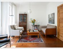 Helles Wohnzimmer mit Holzdielenboden, großem Fenster mit weißen Vorhängen und einem gemusterten Teppich unter einem niedrigen Couchtisch. Im Raum stehen zwei Sessel, ein Sofa, ein Holzvitrinenschrank sowie ein Beistelltisch mit Blumen; an der Decke hängt eine runde Pendelleuchte.