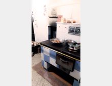 Traditionelle Küche mit einem Ofen im Retro-Stil, bestehend aus blauen und weißen Fliesen. Auf der Ofenplatte stehen Kochgeschirr und Pfannen, im Hintergrund ein eingebauter Holzofen und daneben gestapeltes Brennholz.