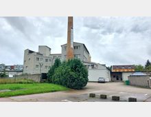 Industriegebäude mit grauer Fassade und einem hohen, gemauerten Schornstein, auf dem das Wort „Luckenwalde“ steht. Im Vordergrund sind eine kleine Grünfläche mit Bäumen, ein Parkplatz und ein angrenzendes Nebengebäude mit gelb-orangefarbener Fassade zu sehen.