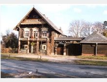 Traditionelles zweistöckiges Holzhaus mit verzierten Balkonen und Dachgiebel, umgeben von ländlicher Umgebung. Rechts davon steht ein kleineres Nebengebäude aus Holz mit Satteldach.