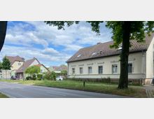 Straße mit freistehenden Häusern, darunter ein weißes Gebäude mit Satteldach und Fensterläden im Mittelpunkt. Im Vordergrund ein Grünstreifen und ein Baum, im Hintergrund weitere Einfamilienhäuser mit roten Dächern unter einem bewölkten Himmel.