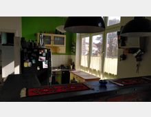 Küche mit grüner Wand, einer Theke aus dunklem Holz und roten Teppichen. Im Hintergrund befindet sich ein großer Kühlschrank, ein Herd mit Dunstabzugshaube, Hängeschränke sowie Fenster mit halbtransparenten Gardinen, die Tageslicht in den Raum lassen.