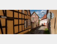 Schmale Gasse mit Kopfsteinpflaster und Fachwerkhäusern in warmen Gelb- und Roséfarben, typisches historisches Straßenbild. Ein Straßenschild mit der Aufschrift „Schloßgasse“ ist links im Bild zu sehen, darüber blauer Himmel mit wenigen Wolken.