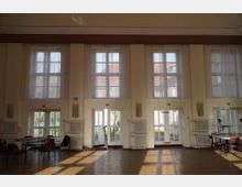 Ein großer Saal mit hohen Decken und großen Fensterfronten, bestehend aus Doppelfenstern und Glastüren, die viel Tageslicht hereinlassen. Der Raum ist schlicht eingerichtet mit einigen Tischen und Stühlen und hat einen hellen Holzboden.