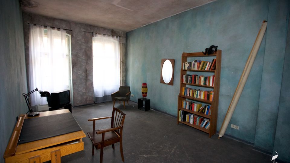 Schlichter Innenraum mit blau gestrichenen Wänden und rohem Boden, zwei hohen Fenstern mit weißen Vorhängen und einem großen Arbeitstisch. An der rechten Wand stehen ein Bücherregal und ein ovaler Spiegel, daneben lehnt ein langes Brett; im Raum stehen zudem ein Sessel und ein Stuhl.