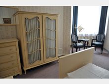 Ein Schlafzimmer mit klassischem Mobiliar, darunter ein cremefarbener Schrank mit Glas- und Zierelementen sowie eine Kommode. Im Hintergrund stehen zwei schwarze Stühle und ein kleiner runder Tisch vor einem großen Fenster mit Vorhängen.