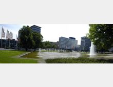 Stadtplatz mit rechteckigem Brunnen und mehreren Wasserfontänen in einer begrünten Umgebung, umgeben von modernen Gebäuden und Fahnenmasten mit verschiedenen Flaggen. Im Hintergrund stehen hohe Bürogebäude, teils aus Glas und Beton.