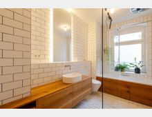 Modernes Badezimmer mit weißen Wand- und Bodenfliesen im U-Bahn-Stil, einem großen Spiegel mit indirekter Beleuchtung, einem Holzwaschtisch mit rundem Aufsatzwaschbecken, einer Badewanne mit Holzverkleidung und einem großen Fenster mit Pflanzen darauf.