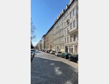 Straßenszene in einer Wohngegend mit Reihen historischer Mehrfamilienhäuser und hellen Fassaden, gepflasterter Fahrbahn, parkenden Autos entlang der Straße und klarem, blauem Himmel. Links sind kahle Bäume sichtbar.