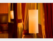 Eine Innenaufnahme zeigt einen warm beleuchteten Raum mit einer schlichten Tischlampe im Vordergrund; dahinter sind rote und beige Vorhänge sowie verschwommene Sitzmöbel zu erkennen.