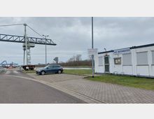 Ein Hafenbereich mit einem metallenen Kran, einem Bürocontainer mit der Aufschrift „HLB Hafenmeister“ und einem geparkten Auto im Vordergrund. Im Hintergrund sind Wasser, Bäume und weitere Hafeninfrastruktur wie ein blauer Hebekran sichtbar.