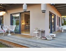 Moderne Terrasse mit Holzboden und überdachtem Bereich, angrenzend an ein Haus mit großen Glasschiebetüren. Auf der Terrasse befinden sich weiße Gartenmöbel, Dekorationen wie hängende Laternen und Pflanzgefäße mit Sukkulenten.