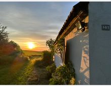 Eine ländliche Szene mit einer kleinen, weißen Hauswand auf der rechten Seite, beleuchtet vom warmen Licht des Sonnenuntergangs. Im Hintergrund sind eine Wiese, Bäume und der weite Himmel sichtbar, während die Sonne tief am Horizont steht.