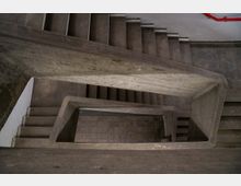 Blick von oben auf ein Treppenhaus aus Beton mit rechteckigem Grundriss und schlichten, grauen Stufen; die Flurebenen sind mit Fliesen ausgelegt, und ein rotes Geländer ist im Hintergrund sichtbar.