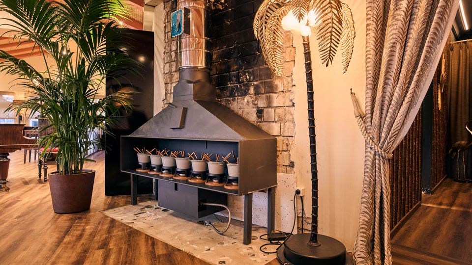 A cozy indoor space featuring a metallic stove with pots arranged in a row underneath, set against a partially charred brick wall. The area is adorned with a potted palm plant, a palm-shaped decorative floor lamp, and patterned curtains, with wooden flooring completing the warm ambiance.
