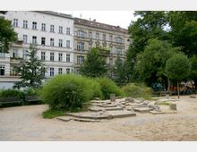 Kleiner Stadtpark mit Sandboden, Bänken und Grünflächen, flankiert von historischen Wohngebäuden im Altbaustil mit dekorativen Fassaden; im Hintergrund zahlreiche Bäume und vereinzelte Personen.
