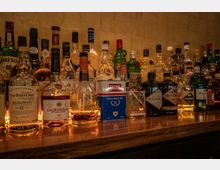 Regal mit einer Auswahl an alkoholischen Getränkeflaschen, darunter Whisky, Cognac, Gin und Spirituosen verschiedener Marken, auf einer dunkelbraunen Holztheke vor einer goldfarbenen Wand arrangiert.