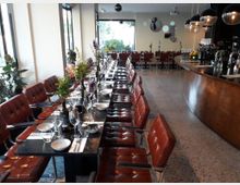 Ein modernes Restaurant mit langen, gedeckten Tischen, umgeben von braunen Lederstühlen. Im Hintergrund befinden sich große Fenster, eine Wanddekoration und eine geschwungene Holztheke mit Hängelampen.