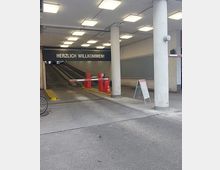 Einfahrt zu einem Parkhaus mit überdachtem Bereich, weißen Säulen und einer Schranke. Über der Einfahrt hängt ein Schild mit der Aufschrift „HERZLICH WILLKOMMEN!“, daneben steht ein Aufsteller.