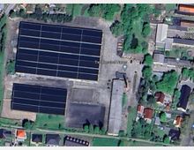 Satellitenansicht eines landwirtschaftlichen Areals, das mehrere große Gebäude mit Solarpaneelen, offene Betonflächen und umliegende Grünflächen zeigt. Im unteren Bereich angrenzend befinden sich Wohnhäuser mit individuellen Grundstücken, und im oberen Bildabschnitt sind Bäume und kleinere Gebäude sichtbar.