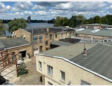 Blick über flache Dächer eines ehemaligen Industrie- oder Gewerbekomplexes mit Backsteinfassaden, Oberlichtern und Innenhof. Im Hintergrund liegen ein breiter Fluss oder See, dichtes Ufergrün und eine entfernte Stadtsilhouette unter wolkigem Himmel.