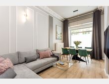 Modernes Wohn- und Esszimmer mit grauem Ecksofa, gläsernem Couchtisch und hellem Holzboden. Im Hintergrund steht ein Esstisch mit grünen Stühlen vor einem großen Fenster mit dunklen Vorhängen; Stuckleisten und Wandpaneele prägen den Raum.