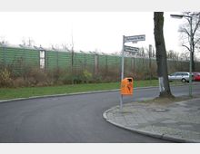 Straßenkreuzung in einem Wohngebiet mit Gehwegen, einem orangefarbenen öffentlichen Mülleimer und Straßenschildern für „Bacharachar Straße“ und „Schaffhausener Straße“. Im Hintergrund verläuft eine grüne Lärmschutzwand, dahinter sind Bäume sichtbar.