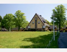 Historisches Gebäude mit gelber Fassade, einem steilen Giebeldach und dekorativen Backsteinakzenten, umgeben von Bäumen und einer großen Wiese. Ein gepflasterter Weg mit Laternen führt am rechten Bildrand entlang.