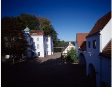 Historischer Hof mit weiß verputzten Gebäuden, roten Ziegeldächern und einem Gebäude mit grünem Kuppeldach; im Hintergrund ein Fachwerkhaus sowie dichte Bäume und ein See. Der Hof ist mit Kopfsteinpflaster bedeckt und von Schatten durchzogen.