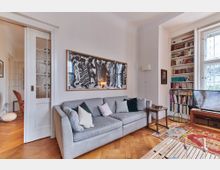 Wohnzimmer mit hellem Holzboden, einer grauen Couch mit Kissen, einem großen Schwarz-Weiß-Kunstwerk an der Wand und einem Bücherregal neben einem Fenster mit verzierten Glasscheiben. Im Raum steht ein Fernseher auf einem Holzschrank, und ein bunter Teppich bedeckt einen Teil des Bodens.