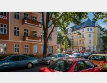 Straßenszene mit Wohnhäusern aus der Gründerzeit, eines in orange-rosé Farben mit weißen Details und eines in gelber Fassade; dazwischen geparkte Autos, Bäume und ein Gehweg.
