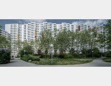 Mehrstöckiges Wohngebäude in moderner Bauweise mit weißer Fassade und farbigen Akzenten, umgeben von einem grünen Park mit Bäumen, Gehwegen und einzelnen Straßenlaternen.