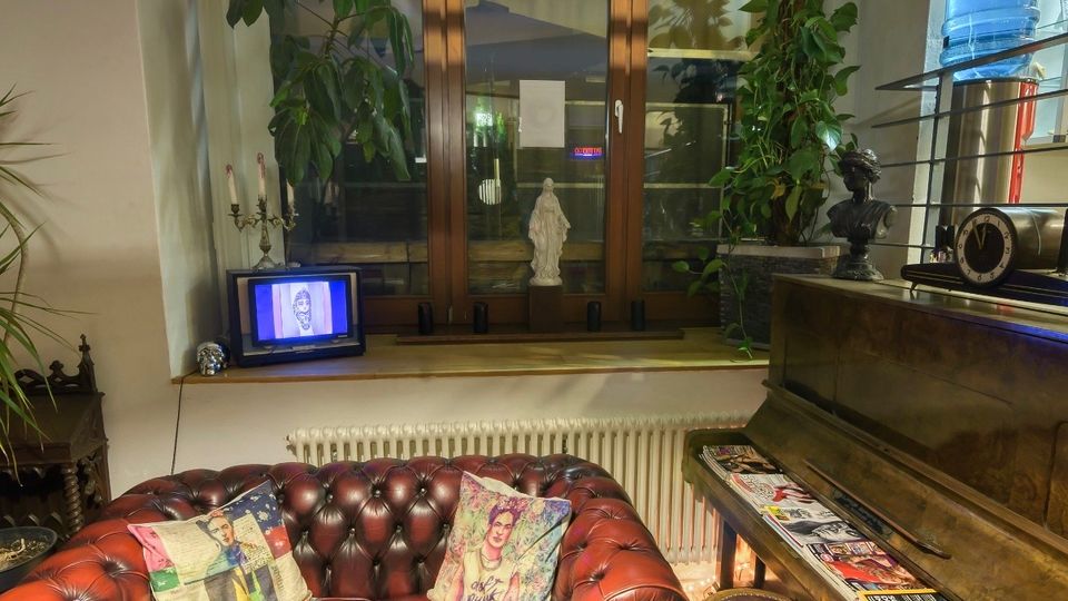 Gemütliche Sitzecke in einem Innenraum mit rotem, geknöpftem Leder-Chesterfield-Sofa vor einem großen Holzrahmenfenster. Daneben stehen ein Klavier mit ausliegenden Zeitschriften, ein kleiner Hocker, Zimmerpflanzen sowie Dekoration auf der Fensterbank.
