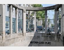 Steinerne Eingangsanlage mit Rundbogen und Säulen zum U-Bahnhof Viktoria-Luise-Platz in Berlin, darunter der Schriftzug „Viktoria-Luise-Platz“. Im Hintergrund sind eine Straße, parkende Fahrräder und Autos sowie mehrere Gebäude zu sehen.