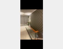 Ein modern gestalteter Flur mit Teppichboden, einer gläsernen Treppenbrüstung mit Holzhandlauf und dekorativer Tapete mit floralen Mustern an der Wand. Im Hintergrund sind geschlossene, weiße Vorhänge und eine Zimmerpflanze sichtbar.