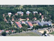 Luftaufnahme eines Wohngebiets mit mehreren gelben Gebäuden mit roten Dächern, die in einer grünen, baumreichen Umgebung angeordnet sind. Im Vordergrund befinden sich Straßen, weitere kleinere Gebäude und freie Flächen, im Hintergrund ein Waldgebiet.