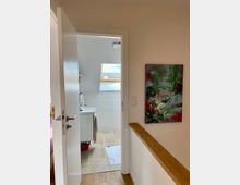 Blick durch eine geöffnete Tür in ein helles Badezimmer mit Fliesenboden, einem Waschbecken und einem Dachfenster, das Tageslicht hereinlässt. Im Vordergrund sind ein Holzboden, eine Wand mit einem abstrakten Gemälde und ein Lichtschalter sichtbar.