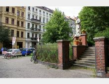Ein kleiner Platz in einer städtischen Umgebung mit gepflastertem Boden, einer Treppe aus Stein und einem Zaun aus Metall und Ziegeln, der zu einer Statue führt. Im Hintergrund sind mehrstöckige Wohngebäude mit verzierten Fassaden, Balkonen und parkenden Autos zu sehen, umgeben von Bäumen.