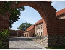 Blick durch einen gemauerten Rundbogen aus roten Ziegelsteinen auf einen Innenhof mit Kopfsteinpflaster. Umgeben sind der Hof und ein Gebäude im Hintergrund von historischen Ziegelsteinmauern mit roten Dächern, teils von Bäumen beschattet.