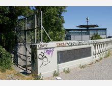 Brückengeländer der Bösebrücke in Berlin, dessen Steinsockel mit Graffiti bemalt ist, daneben ein schmiedeeisernes Tor. Im Hintergrund ist ein modernes S-Bahn-Gebäude mit Glasfassade und S-Bahn-Logo zu sehen.