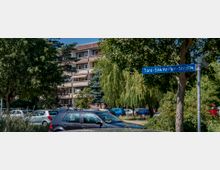 Mehrstöckiges Wohngebäude mit Balkonen im Hintergrund, umgeben von Bäumen und geparkten Autos. Im Vordergrund ein blauer Straßenschild mit der Aufschrift „Toni-Stemmler-Straße“.