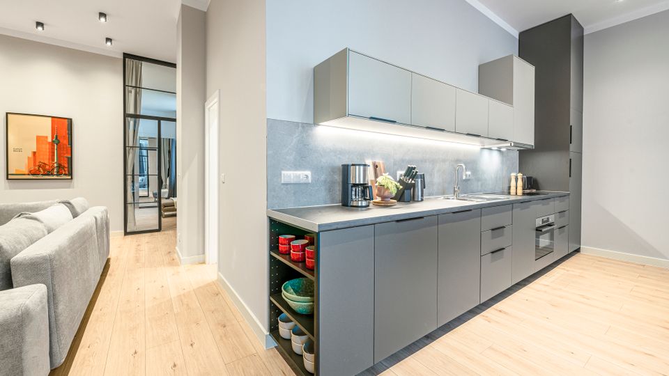 Moderne, offene Wohnküche mit langer, grauer Einbauzeile, Oberschränken und integrierten Geräten, beleuchtet durch Einbauspots und Unterbauleuchten. Heller Holzboden und angrenzender Wohnbereich mit grauem Sofa sowie Glastür zum Flur sind sichtbar.