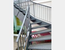 Schwarze Wendeltreppe aus Metall mit rutschfester Oberfläche vor einer hellen Gebäudefassade. Im Hintergrund befinden sich eine verglaste Tür und rote Gartenmöbel auf einer Holzterrasse.