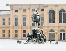 Reiterdenkmal vor einem historischen gelben Gebäudefassade mit hohen Fenstern und dekorativen Pilastern; der Schnee bedeckt den Boden und die Statue.