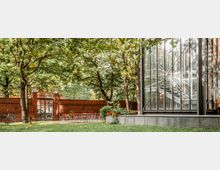 Ein Garten mit mehreren Tischen und Stühlen auf einem gepflasterten Bereich, umgeben von hohen Bäumen und einer roten Ziegelmauer. Rechts im Bild steht ein modernes Gebäude mit einer gläsernen Fassade und sichtbaren Treppen im Inneren.