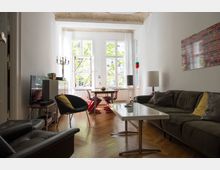 Wohnzimmer mit Holzboden und großen Fenstern, durch die Tageslicht hereinfällt. Es ist möbliert mit einer dunkelgrauen Couch, mehreren Sesseln, einem weißen rechteckigen Couchtisch, und einem Esstisch mit roten Stühlen; an der Wand hängt ein farbiges Wandbild.