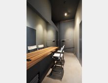 Ein schmaler Raum mit einem langen Holztisch, darauf zwei Klappstühle mit heller Bespannung. Der Raum hat eine graue Wandgestaltung, einen Spiegel an der linken Wand und eine Kleiderstange mit Bügeln im Hintergrund. Eine einzelne Deckenleuchte beleuchtet den Raum.