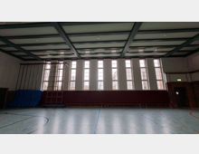 Innenansicht einer Sporthalle mit hohem Rasterdecken-Tragwerk, großer Fensterfront aus schmalen, hohen Fenstern und markierten Hallenbodenlinien. An der Wand stehen Kletterstangen und eine Sprossenwand mit blauen Turnmatten; rechts ist ein Ausgang in einer Ziegelwand.