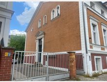 Ziegelgebäude mit orange-brauner Fassade, weißen Fenstereinfassungen und einem Haupteingang mit einem kleinen Vordach. Im Vordergrund ist ein Metalltor mit Ziegelpfeilern zu sehen, das einen gepflasterten Hofbereich abgrenzt.